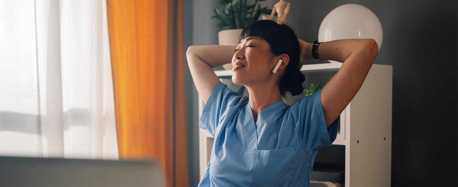 A nurse in blue scrubs takes a mindful break, relaxing in a chair with earbuds in, illustrating the importance of sustainable employee wellness for preventing burnout.
